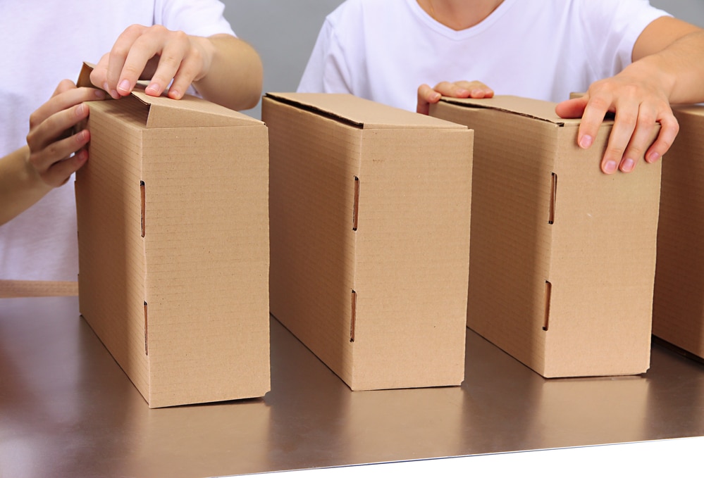 Two people in white shirts sealing cardboard boxes on a table, preparing packages for shipping or storage.