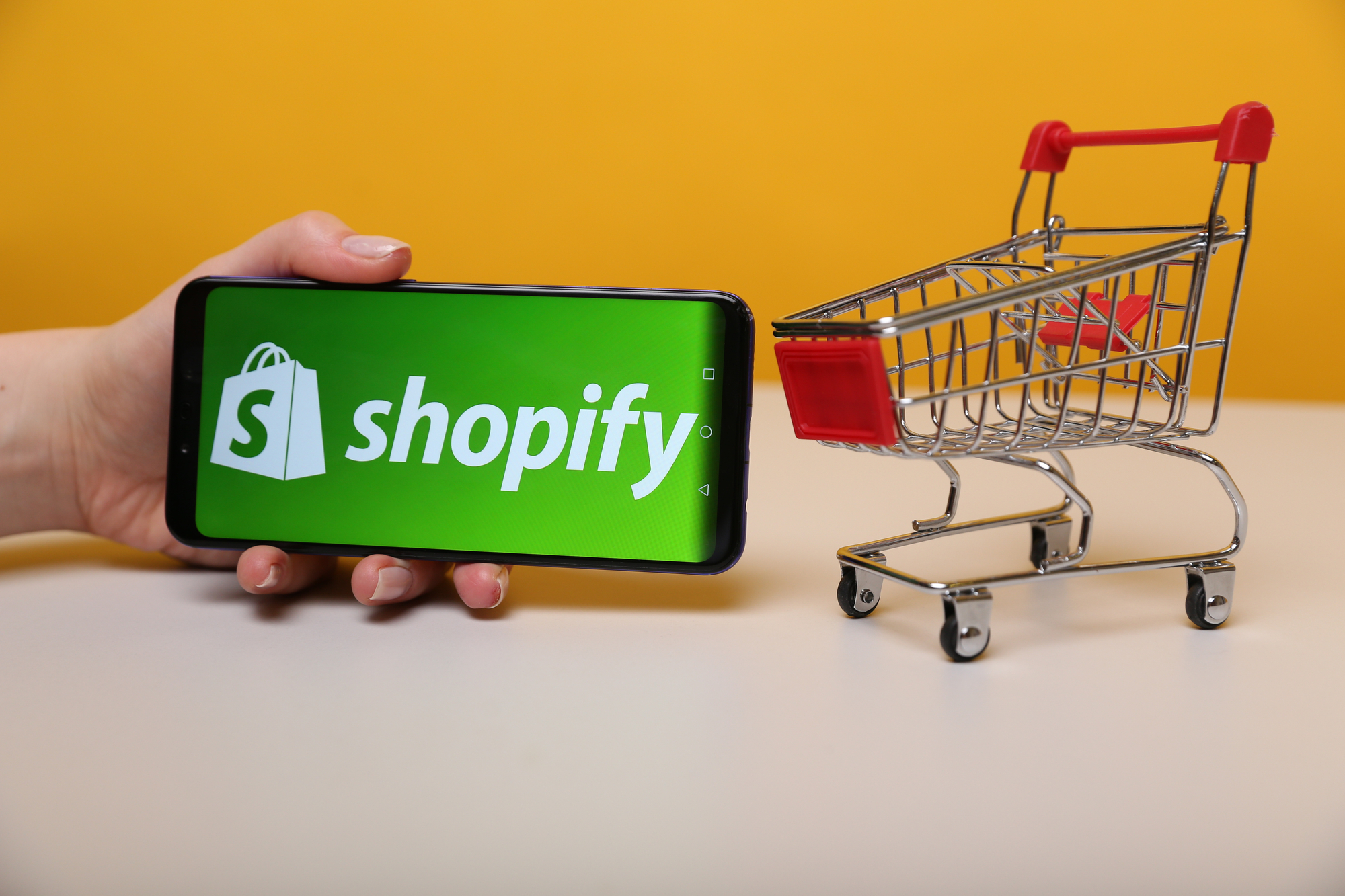 Shopify logo on smartphone beside miniature shopping cart