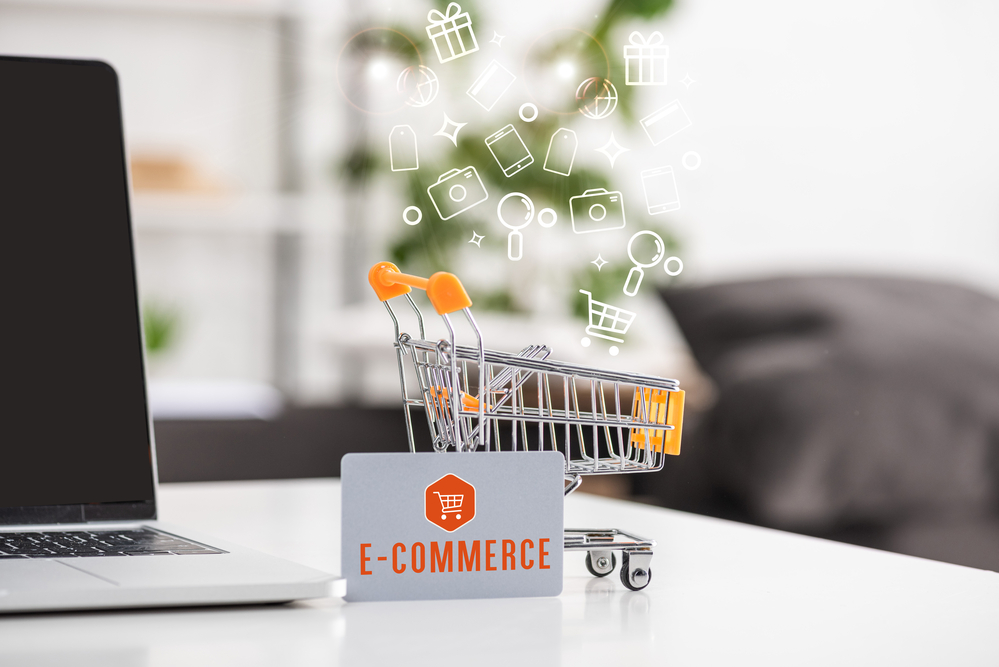 miniature shopping card beside a laptop on a desk with a placard saying ecommerce