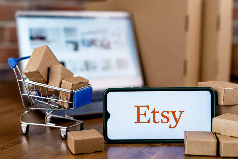 Etsy logo on a mobile phone in front of an open laptop with a miniature full shopping cart beside it.